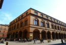A large historic building with arched windows and colonnades, located in a European city square, with people walking and gathering around under a clear blue sky.