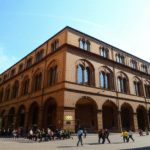 A large historic building with arched windows and colonnades, located in a European city square, with people walking and gathering around under a clear blue sky.